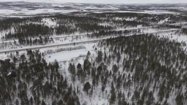 İHA görüntülerinde geniş kar örtülü manzaralar, yoğun ormanlar ve İsveç 'in Kiruna kenti yakınlarında asfalt bir otoyol görülüyor ve yukarıdan gerçek zamanlı bir kış manzarası görülüyor..