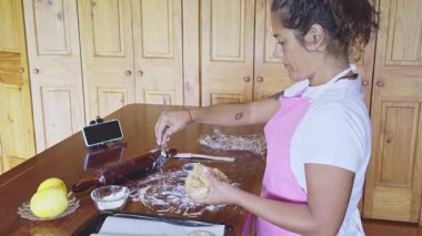 Pembe önlüklü bir kadın rahat bir mutfak ortamında ustalıkla kurabiye hazırlıyor. Malzemeler ve merdaneler ahşap masada görülebiliyor, sıcak ve rahat bir atmosfer yaratıyor..