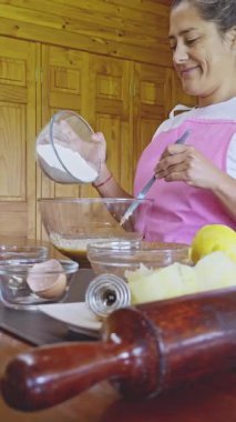 Pembe önlüklü kadın bir kaseye un döker, sıfırdan kurabiye pişirmeye hazırlanır. Tezgahta yumurta ve limon gibi temel pişirme araçları ve taze malzemeler görülüyor.