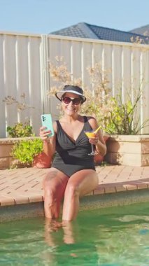 Siyah mayo giymiş bir kadın havuz başında oturmuş, ferahlatıcı bir içki ve güneş ışığının tadını çıkarıyor. Rahatlamış görünüyor, güneşli bir günün sakin havasını yakalıyor.