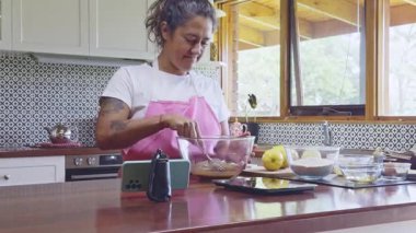 Bir kadın, modern bir mutfakta telefonundaki kurabiye tarifini takip ederken cam bir kasede malzemeleri karıştırıyor. Taze malzemeler ve çatallar tezgahın üstüne yerleştirilmiş.