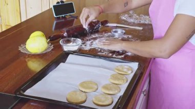 Pembe önlüklü bir kadın ahşap bir masada ev yapımı kurabiyeler hazırlıyor. Hamuru düzleştirmek için merdane kullanıyor, yanında limon ve akıllı telefon var, pişirmeye hazır.