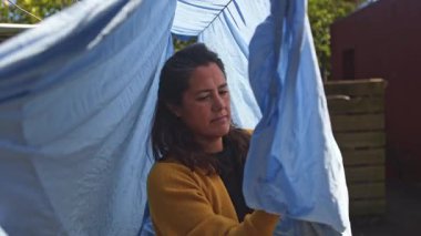 Güneşli bir gökyüzünün altına mavi bir çarşaf asan bir kadın, günlük rutin bir iş yapıyor, açık hava ortamında odaklanma ve kendini adama, günlük hayatı gözler önüne serme.