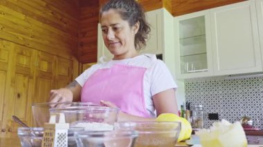 Pembe önlük giyen bir kadın sıcak mutfağında kurabiye pişirir. Etrafı fırın aletleri ve taze limonlarla çevrili cam bir kasede malzemeleri karıştırıyor.