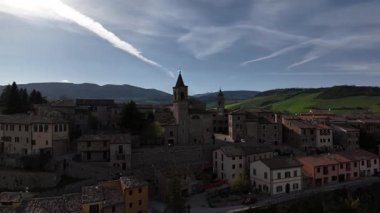 İtalya 'nın Belforte del Chienti kentinin insansız hava aracı görüntüsü, taş binaların üzerinde sıcak günbatımı ışığı, renkli evler ve arka planda yuvarlanan yeşil tepeler.