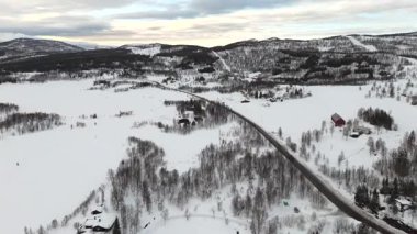 Norveç 'te sakin bir Nordik kış manzarasının havadan görünüşü, donmuş bir nehir ve bir dron tarafından yakalanan dolambaçlı asfalt bir yol içeriyor. Karla kaplı arazi seyrek bitki örtüsü ve ıssız binalarla dolu..