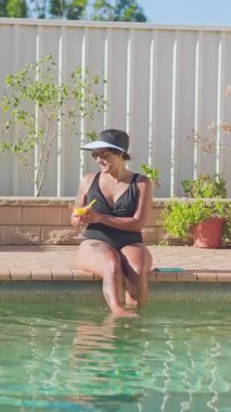 Güneşli bir günde, siyah mayo ve güneşlik takan bir kadın havuz kenarında dinlenir. Ayaklarını berrak suya daldırıp ferahlatıcı bir kokteylin tadını çıkarıyor.