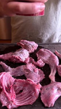 Koyu ahşap kesme tahtasındaki çiğ et parçalarını tuz ve biberle tatlandıran bir adamın yakın çekimi. Sahne, kırsal bir mutfak ortamında ev yemeği hazırlığında bir basamak yakalıyor.