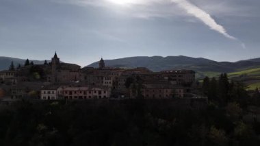 İtalya 'nın Belforte del Chienti kentinin insansız hava aracı görüntüsü, taş binaların üzerinde sıcak günbatımı ışığı, renkli evler ve arka planda yuvarlanan yeşil tepeler.