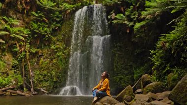 Sarı ceketli bir kadın Avustralya 'nın Büyük Okyanus Yolu' nun yemyeşil şelalesinde kayaların üzerinde oturmuş manzaranın huzurlu doğal güzelliğinin tadını çıkarıyor..