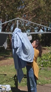 Bir kadın açık gökyüzünün altında arka bahçesindeki bir çamaşır ipine mavi bir çarşaf asıyor. Güneş ışığı ağaçların arasından süzülüp sakin bir atmosfer yaratır.