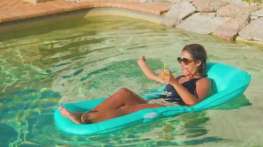 Bir kadın, elinde kokteyl tutarak, mavi bir havuz kenarında güneşli bir günün tadını çıkarır. Güneş gözlüğü ve mayo takıyor. Etrafı kristal berraklığında su ve rahatlama ile çevrili.