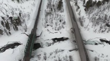 İsveç 'in Kiruna kenti yakınlarındaki bir yol ve tren yolu ile kesişen karlı araziyi gösteren İHA görüntüleri. Video, dondurulmuş ağaçlar ve kısmen donmuş nehirlerle dolu bir kış sahnesini gerçek zamanlı olarak kaydetti..