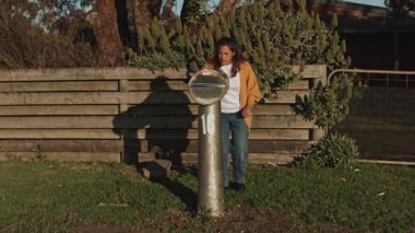 Sarı ceketli bir kadın evinin dışındaki posta kutusunu kontrol ediyor. Etrafı yeşillik ve ahşap çitlerle çevrili. Bu görüntü, banliyö ortamındaki basit ve günlük bir rutini yakalar.