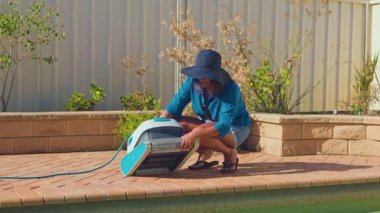 Güneşli bir günde güneş şapkası takan ve günlük giysiler giyen bir kadın, bir robotik havuz temizleyicisinin altını kontrol eder, iyi korunmuş bir Avustralya arka bahçesinde havuz bakımı görevlerini yerine getirir.