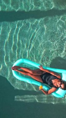 Siyah mayo giymiş bir kadın şişme bir havuzda güneşli günün tadını çıkarıyor. Elinde ferahlatıcı bir içecek tutuyor, boş zamanlarını ve yaz havasını somutlaştırıyor.