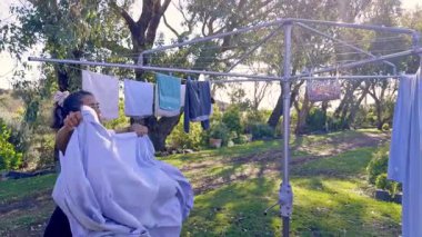 Bir kadın açık havada çamaşır ipinde yeni kurumuş çamaşır topluyor. Güneş ışığı yakındaki ağaçların arasından süzülür, taze kesilmiş çimenlerin üzerine gölgeler düşer, dingin bir atmosfer yaratır.