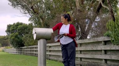Kırmızı kazak giyen bir kadın posta kutusunu ahşap bir arka planla kontrol ediyor. Bu görüntü, kırsal kesimdeki günlük rutinlerden bir anı yakalar.