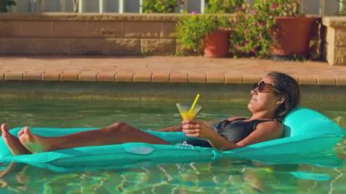 Mavi şişme bir şezlongda, sıcak güneşin altında kokteyl içen bir kadın. Rahatlama, huzur ve yaz titreşimleri bu huzurlu anı tanımlar.