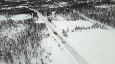 Drone, Norveç 'te karla kaplı ormanlarla çevrili asfalt bir yolda seyahat eden bir minibüsü görüntüleyerek, sakin kış manzarasını ve yol koşullarını vurguladı..
