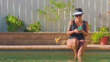 Bir kadın havuz kenarında, ayakları suyun içinde oturarak rahatlıyor. Güneş gözlüğü ve güneşlik takıyor, içkinin tadını çıkarıyor ve akıllı telefonunu kullanıyor.