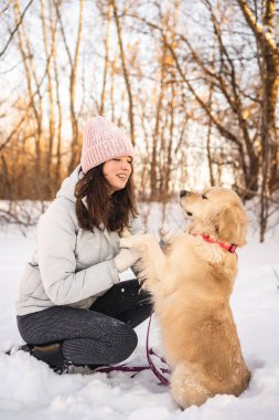 Bereli ve kışlık ceketli bir kadın karda diz çöker, karlı bir ormanda Golden Retriever ile neşe içinde karşılaşır, samimi bir arkadaşlık sahnesi yaratır..