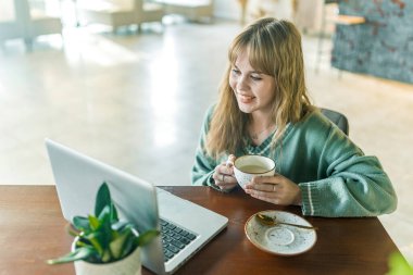 Sarışın bir kız, rahat bir kahve dükkanında, tahta bir masada dizüstü bilgisayar ve küçük bir bitkiyle oturup, huzurlu ve rahat bir atmosferi yakalıyor.