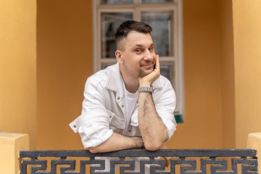 Beyaz ceketli, gülümseyen mutlu bir işadamı, düşünceli bir ifadeyle balkonda rahatlarken, kentsel bej bir binanın arka planına çevrilmiş, kameraya bakarken görülüyor..