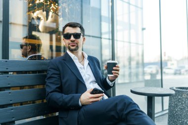 Koyu takım elbiseli ve güneş gözlüklü kendine güvenen bir işadamı açık havada kahve molası verir. Bir şehir ortamında modern profesyonellik yayarak akıllı bir telefon tutuyor.