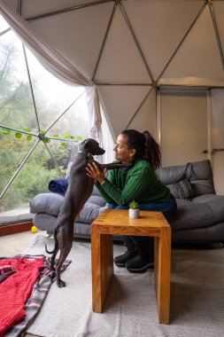 Yeşil kazaklı bir kadın rahat bir glamping içinde köpeğiyle neşe içinde etkileşime geçiyor. Kurulumda rahat bir kanepe, ahşap masa ve battaniyeler yer alıyor..