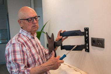 Yaşlı bir adam, bir tornavida kullanarak bir televizyon duvarı montajı DIY kurulumuna odaklanır ve parantezi dikkatle inceler.