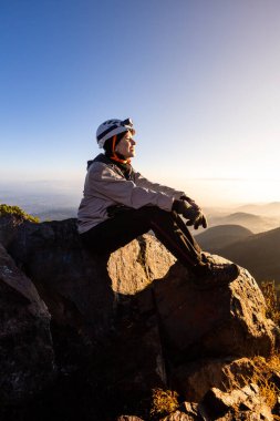 Açık hava teçhizatı ve kasklı bir yürüyüşçü bir kayanın üzerinde oturur ve dağın zirvesinden güneşin doğuşunun tadını çıkarır. Güneş dingin manzaranın üzerine ılık bir parıltı saçar..