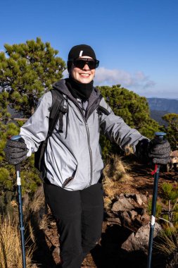 Güneş gözlüğü ve kış giysileri takan neşeli bir yürüyüşçü yeşil çam ağaçları ve açık mavi gökyüzü ile çevrili güneşli bir dağ yolunda yürüyüş direkleri ile ayakta duruyor..
