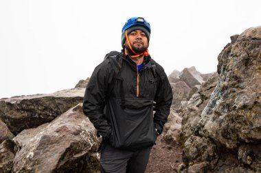 Yağmurluk ve güvenlik kaskı takan kararlı bir yürüyüşçü kayalık bir dağ yolunda güven içinde duruyor. Sisli atmosfer macera havasını arttırır..