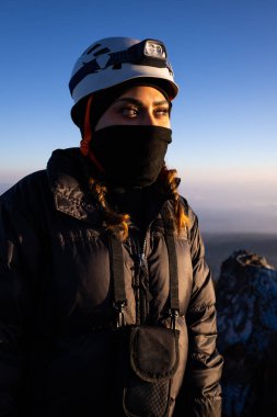 Kask, far ve kalın koruyucu elbiseler giyen bir kadın dağa tırmanmaya hazırdır. Sabahın erken saatlerinde sakin bir parıltı yayar, kararlılıklarını vurgular..