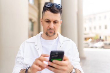 Sıradan beyaz kıyafetler giymiş odaklanmış bir erkek işadamı kalabalık bir caddede akıllı telefon kullanıyor..