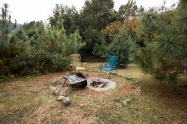 İki renkli metal sandalye ve glamping ile çevrili bir ateş çukurunun sakin bir açık hava sahnesi. Yeşillik ve çam ağaçları rahatlatıcı bir bahçe atmosferi yaratır..