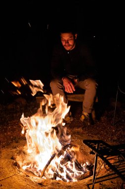 Bir insan, gece karanlığında yanan bir kamp ateşinin yanında sessizce oturur. Sıcak parıltı çevreyi aydınlatıyor, açık havada sakin ve düşünceli bir atmosfer yaratıyor..