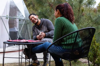 İki insan dışarıda canlı bir sohbetin tadını çıkarıyorlar, rahat bir bahçe alanında, göz alıcı bir kubbenin yanında oturuyorlar. Rahatlamış ve neşeli görünüyorlar, doğayla çevrili..