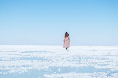 Paltolu bir kadın, açık mavi bir gökyüzünün altında donmuş gölde yürüyor. Huzurlu sahne, doğada bir sükunet ve yalnızlık hissi yakalıyor..