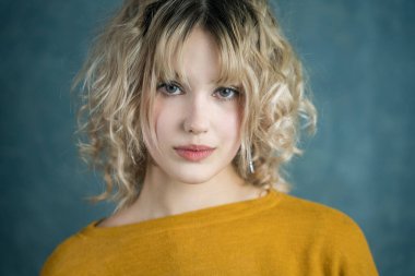 Sarı kazak giyen, dalgalı sarı saçlı genç bir kadının yakın portresi. Özgüvenle kameraya bakıyor, dokulu mavi bir arka planda, sanatsal ve çarpıcı bir görüntü yaratıyor..