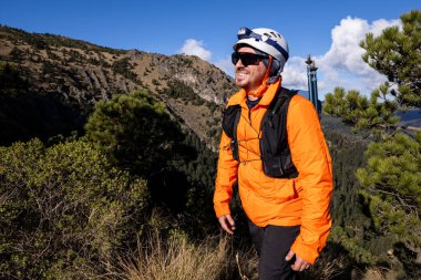 Turuncu ceketli, kasklı, güneş gözlüklü, güneşli bir günde dağlık araziyi keşfeden gülümseyen bir maceraperest. Sahne, açık hava macerasının neşe ve başarısını yakalar..