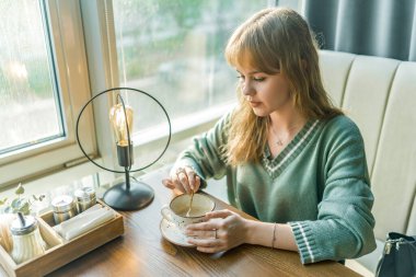 Genç sarışın bir kız, rahat bir kahve dükkanında huzurlu bir anın tadını çıkarıyor. İçkisini düşünceli bir şekilde karıştırıyor. Yumuşak bir ışık ve sıcak bir atmosfer rahatlama için sakin bir ortam yaratır.