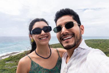 Neşeli bir erkek ve kadın Cozumel manzarasının önünde selfie çekiyorlar yemyeşil ve okyanus manzarası sergiliyorlar. İkisi de güneş gözlüğü takıyor ve gülümsüyor. Birlikte maceralarının bir anını yakalıyorlar..