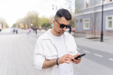 Şık beyaz elbiseli genç erkek işadamı, modern teknoloji kullanımı ve kentsel yaşam tarzını yansıtan hareketli bir şehir caddesinde akıllı telefon kullanıyor..