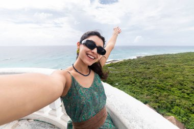 Neşeli bir kadın Cozumel, Meksika 'da manzaralı bir manzarada selfie çekerken sevinçle kolunu kaldırıyor. Güneş gözlüğü takıyor, desenli bir elbise ve bereketli kıyı manzarasıyla çevrili..