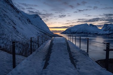 Huzurlu bir kış sahnesi, Norveç 'in Senja kentindeki bir yolculukta, görkemli dağlarla kaplı bir yolun üzerinde gün batımını ve uzakta sakin bir fiyordu yakalar..
