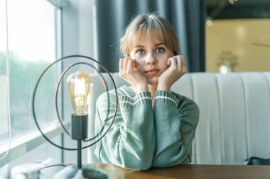 Sarışın bir kız, rahat bir kahve dükkanında nazikçe oturup çenesini ellerinin üzerinde dinlendiriyor. Yumuşak bir ışık ve sıcak bir atmosfer huzurlu anı için sakin bir zemin oluşturur.