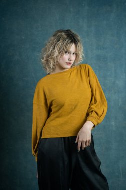 Genç bir kadın, mavi desenli bir arka fona karşı sarı bir kazak sergiliyor. Gelişigüzel ama şık pozlar moda ve kişiliği sıcak bir ortamda ön plana çıkarıyor..