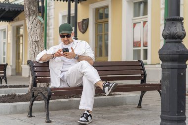 Beyaz giyinmiş bir erkek işadamı şehir bankında oturuyor, akıllı telefon kullanıyor, sakin bir şehir zeminine sahip..